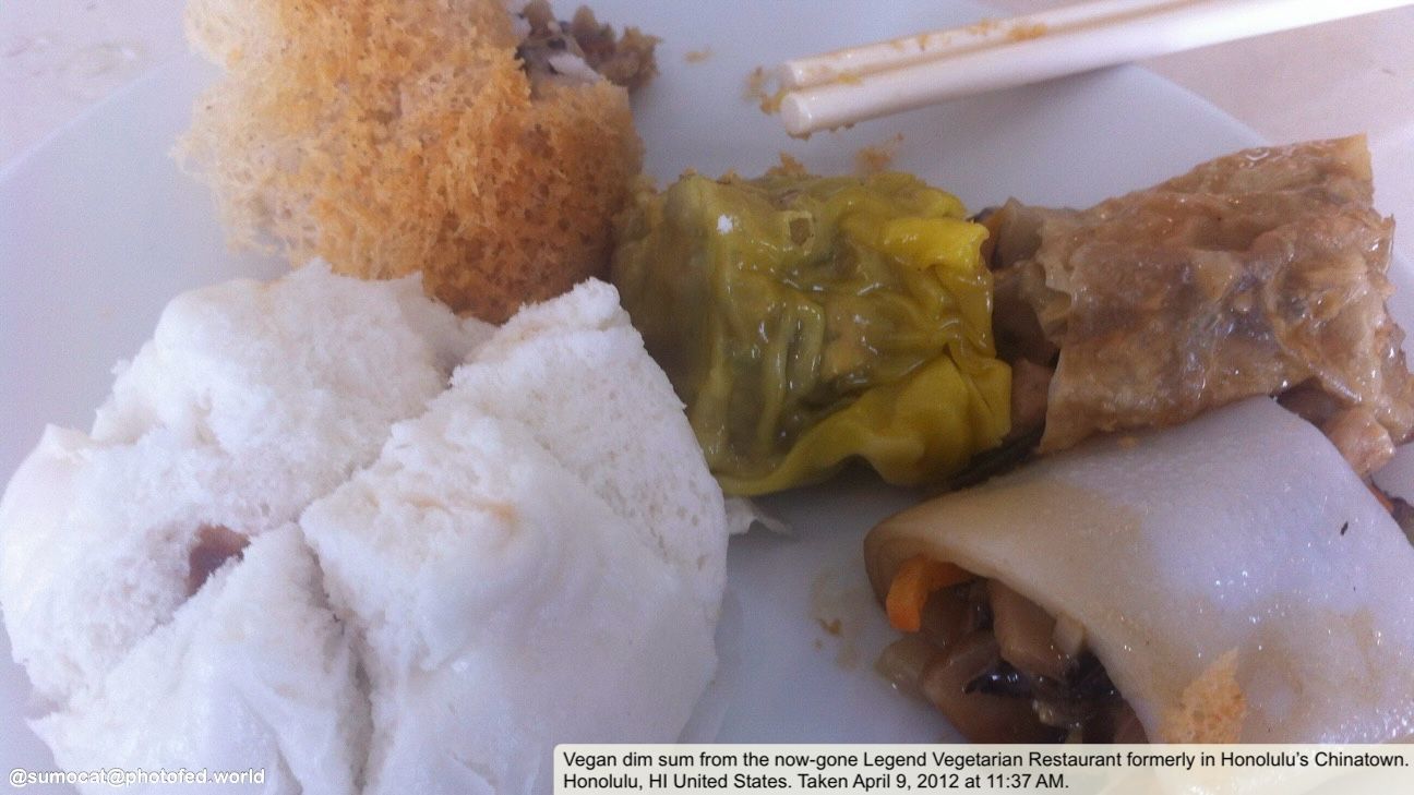 Selection of vegan dim sum, including “char siu” bun, taro dumpling, flat noodle roll filled with vegetables, and shu mai, on a white plate with round chopsticks hanging off the side.