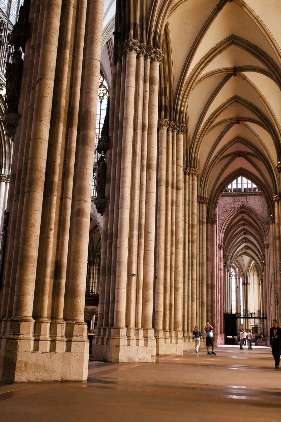 A view along a side aisle in the interior of the Cologne Cathedral, focusing on the columns along the central nave.
The people in the distance look tiny compared to the columns and the ceilings at 18m high.
The stone is light and shifts to a pink hue towards the East end, as its illuminated by stained glass windows (out of view).